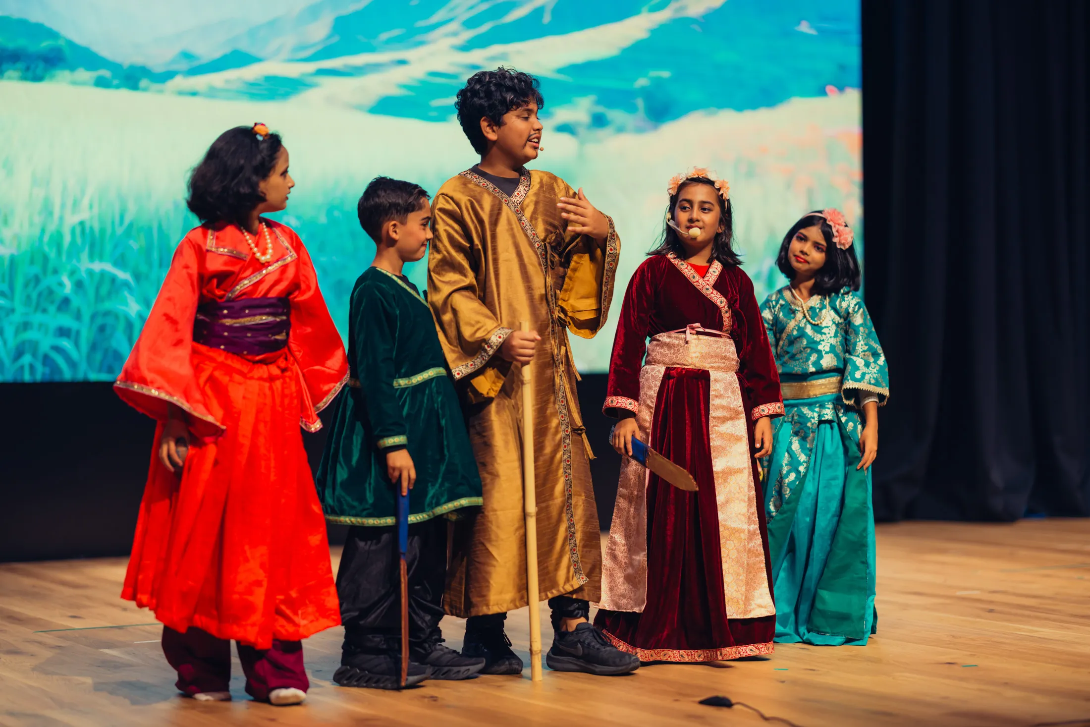 Five children in colorful traditional costumes performing on stage with microphones and props.