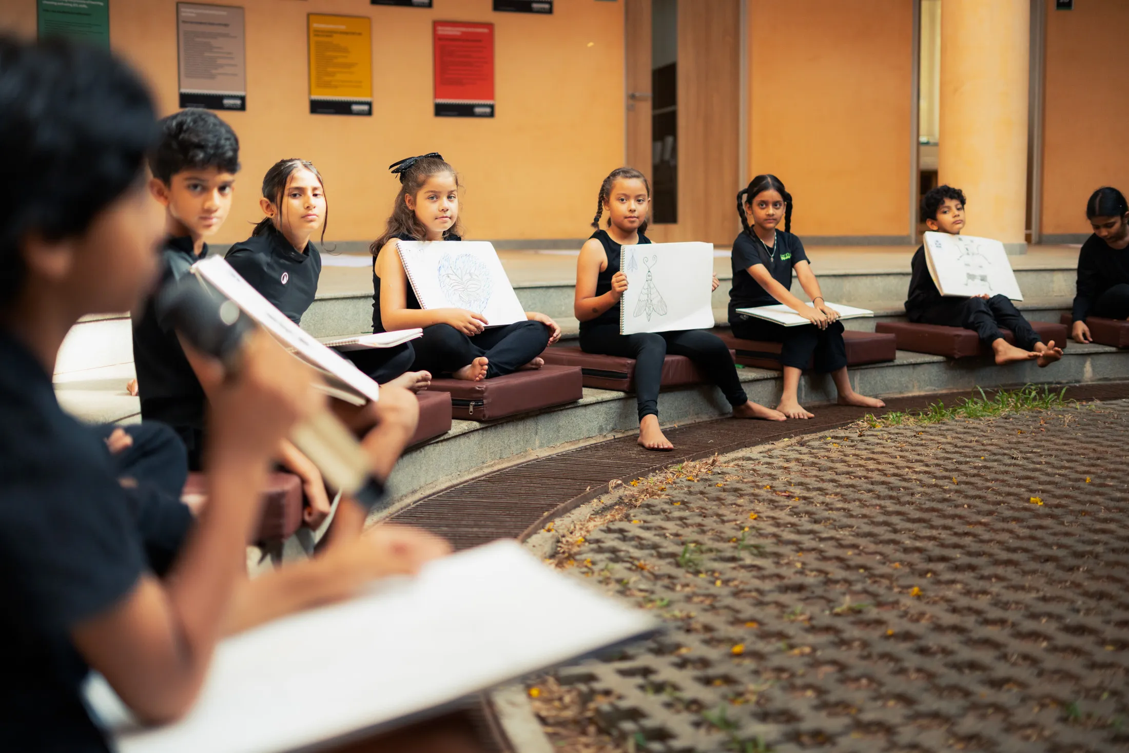 Children seated in a semicircle holding and showing their sketchbook drawings in an outdoor area.