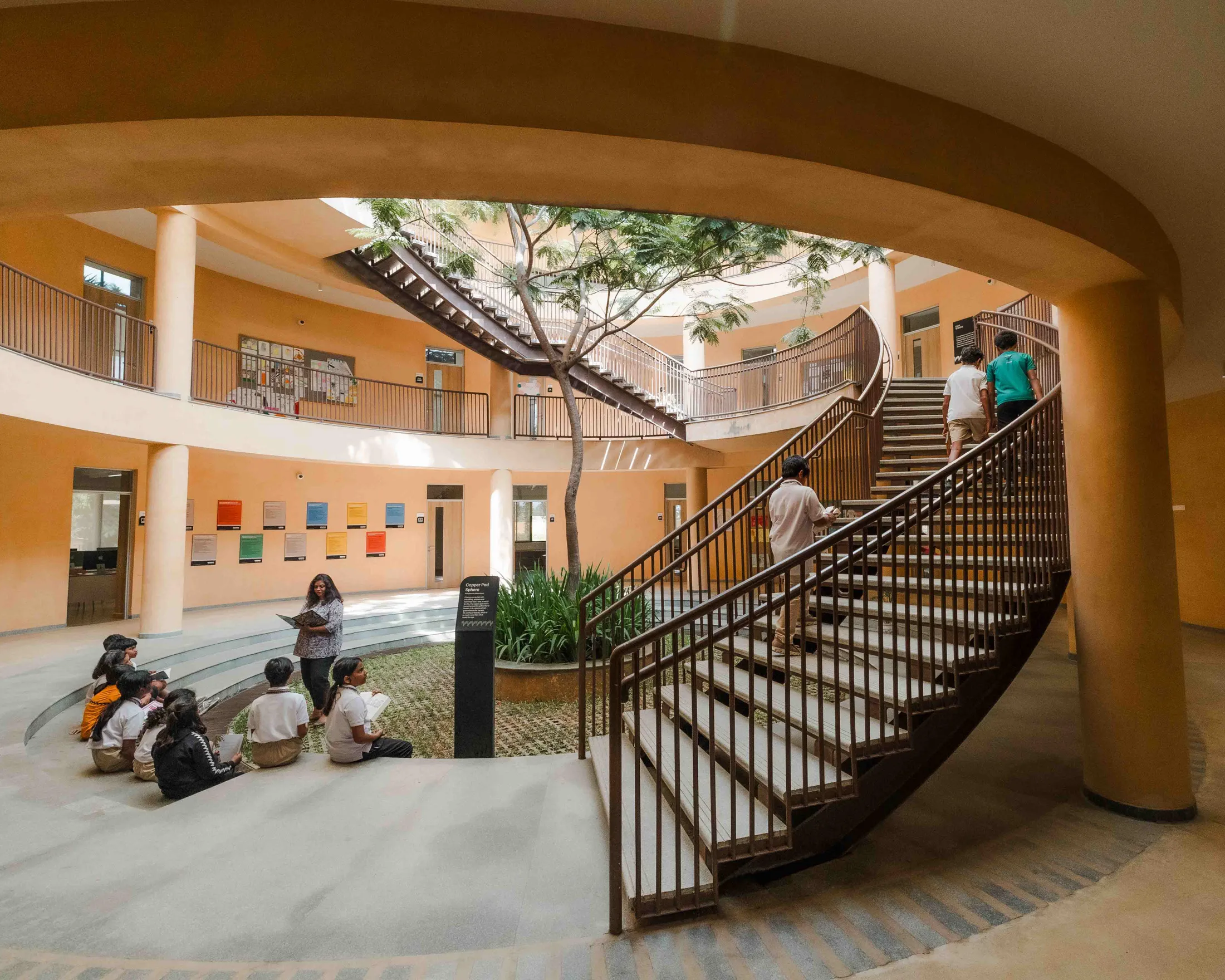 Indoor circular courtyard with a tree, staircase, and students sitting and walking inside a school building.