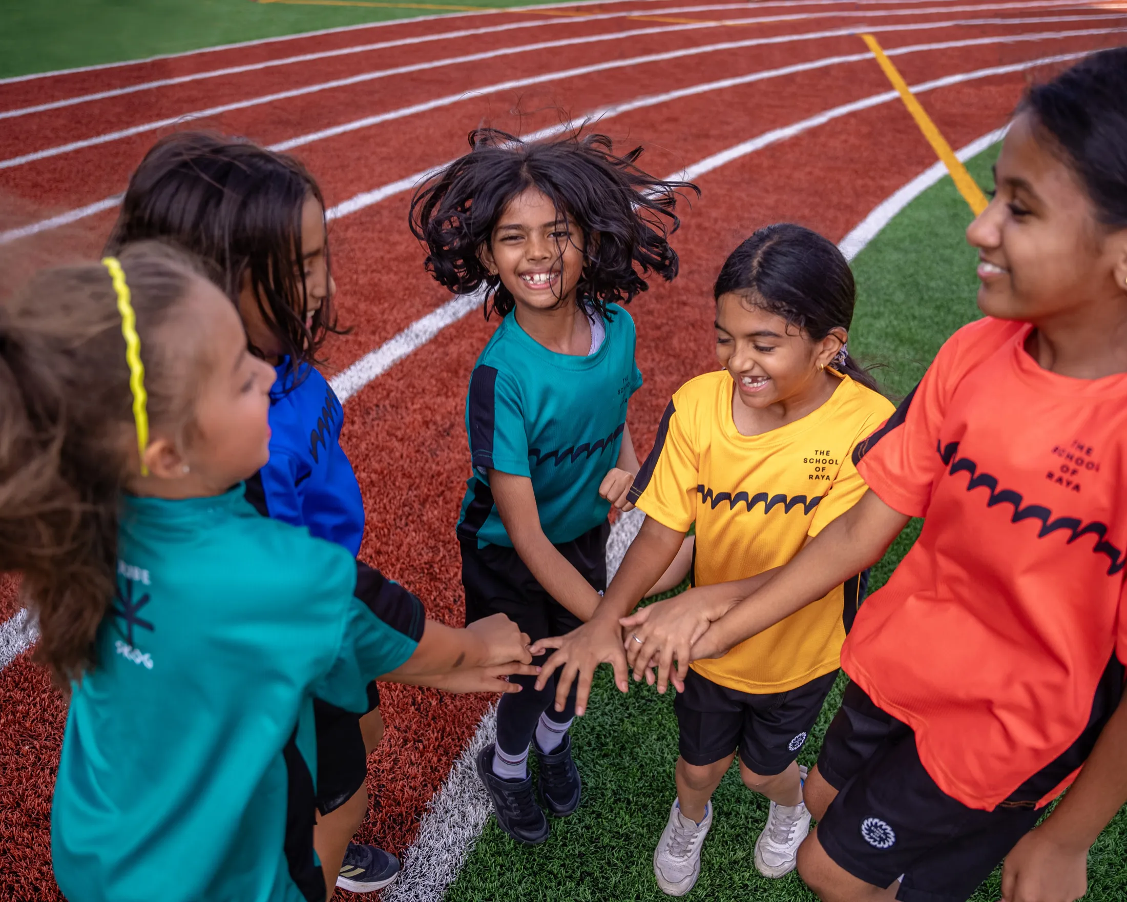Five kids in colorful sports shirts joining hands on running track, smiling and laughing.