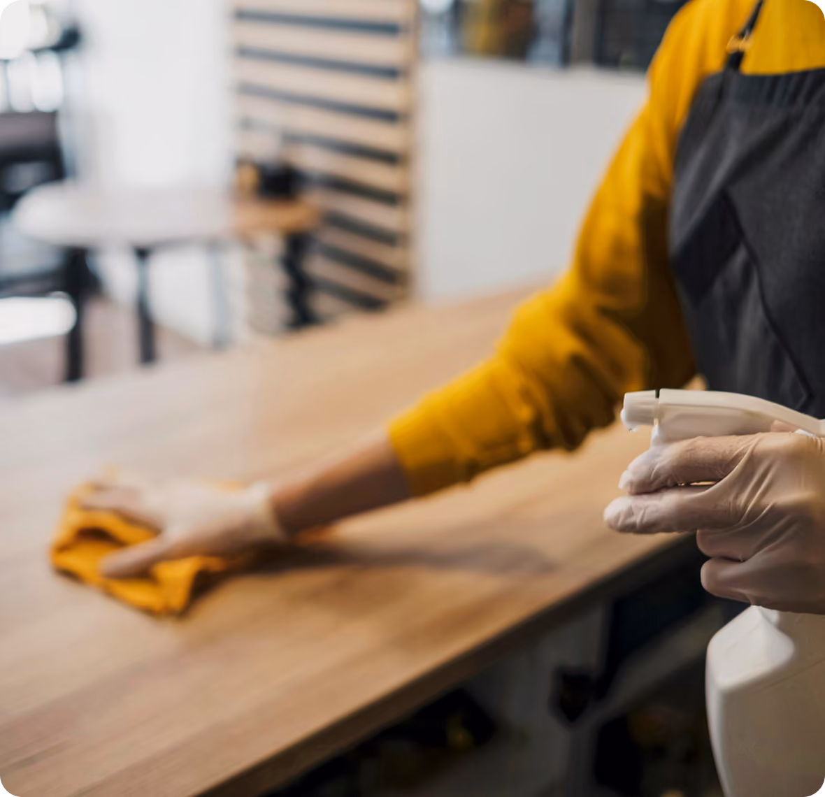 Person mit Reinigungshandschuhen wischt einen Holztisch mit einem Tuch ab und hält eine Sprühflasche mit Reinigungsmittel in der Hand.