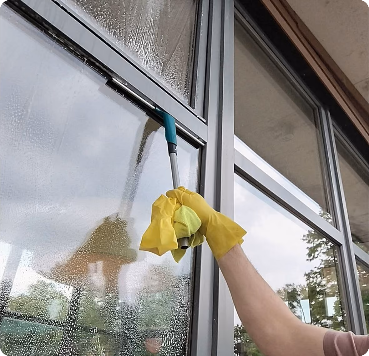 Person in gelben Reinigungshandschuhen reinigt ein großes Fenster im Außenbereich mit einem Fensterabzieher.