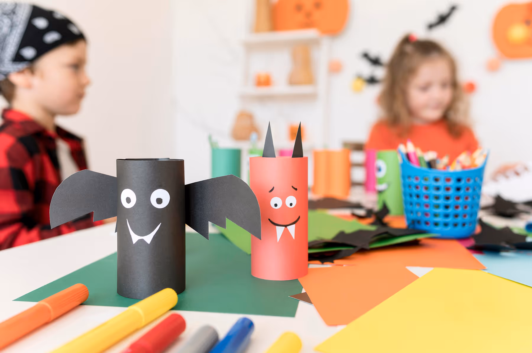 Two Halloween-themed crafts made from colored paper rolls, one designed as a black bat and the other as a red devil, with children and craft supplies in the background.