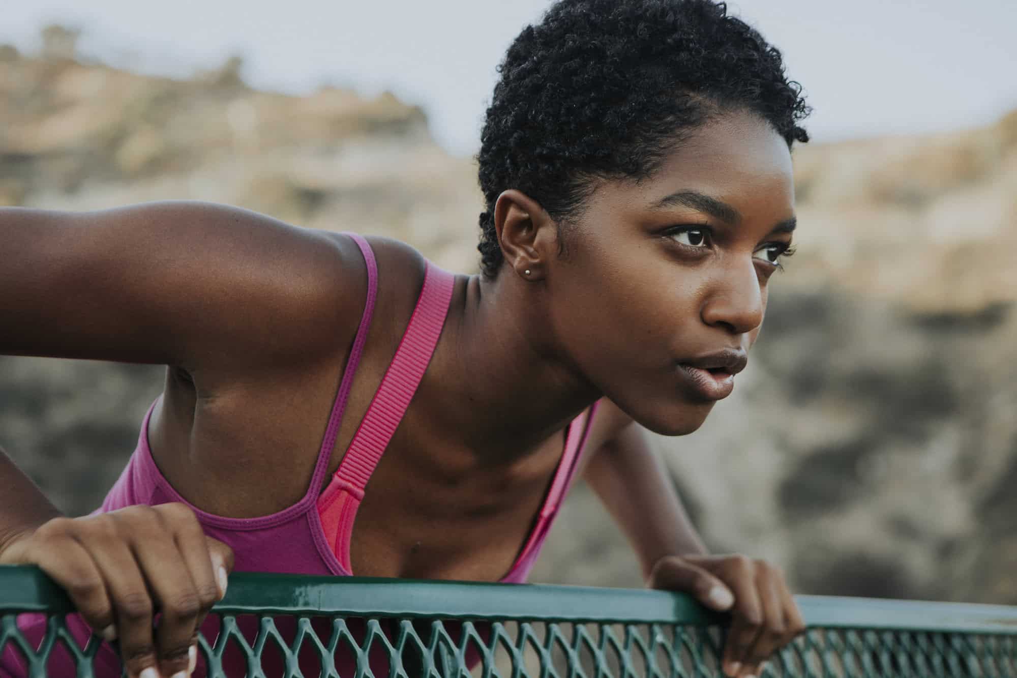 Close-up of a woman in a pink top leaning on a green fence outdoors, focused and determined.