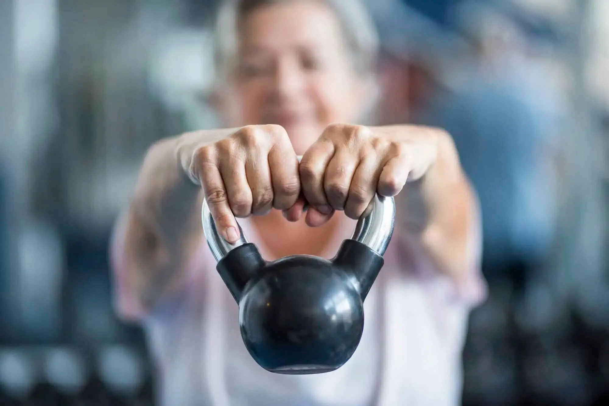 Senior person holding a black kettlebell with both hands, stretching arms forward in a gym.