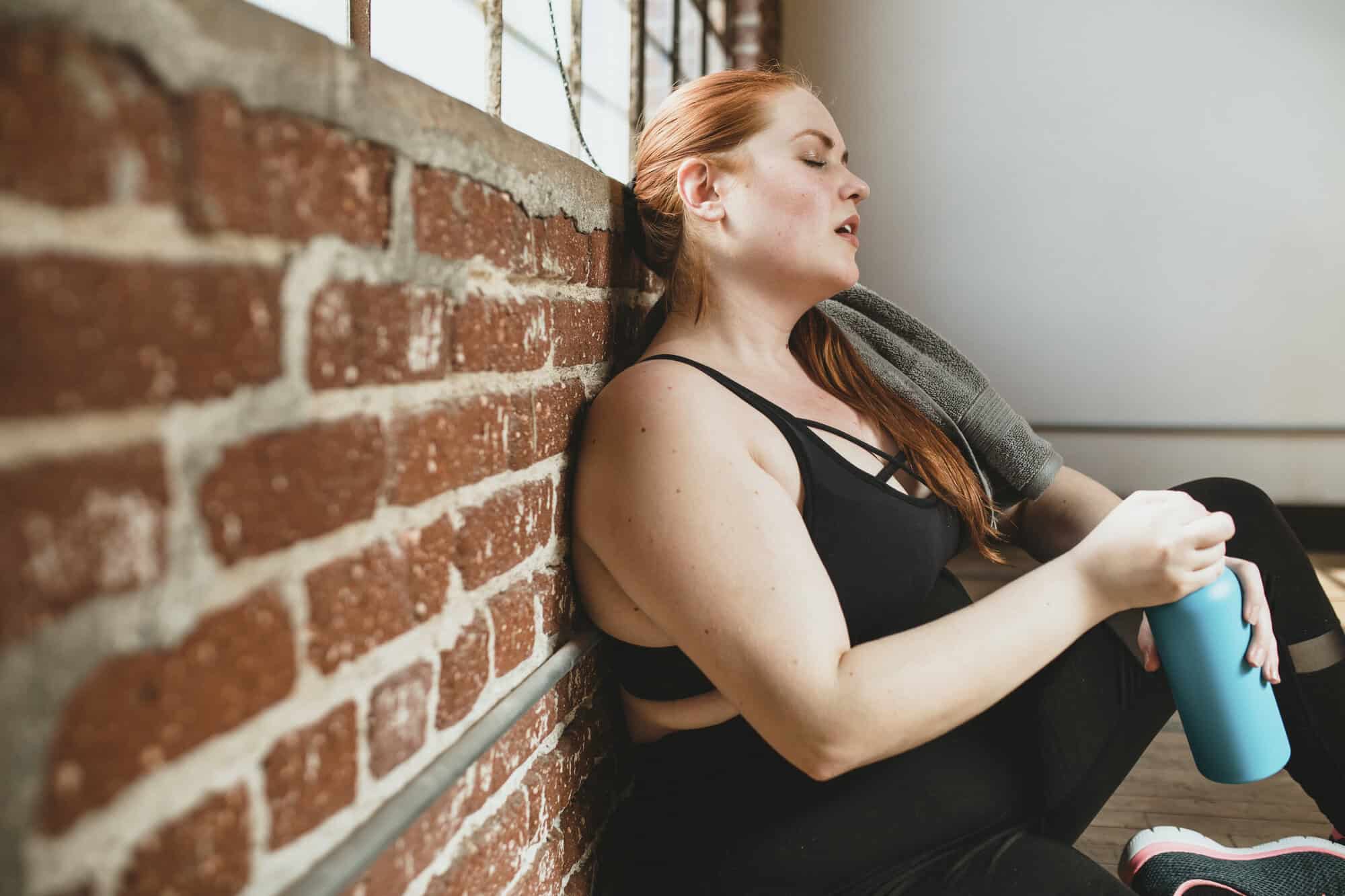 Woman in black workout clothes resting against a brick wall holding a blue water bottle.