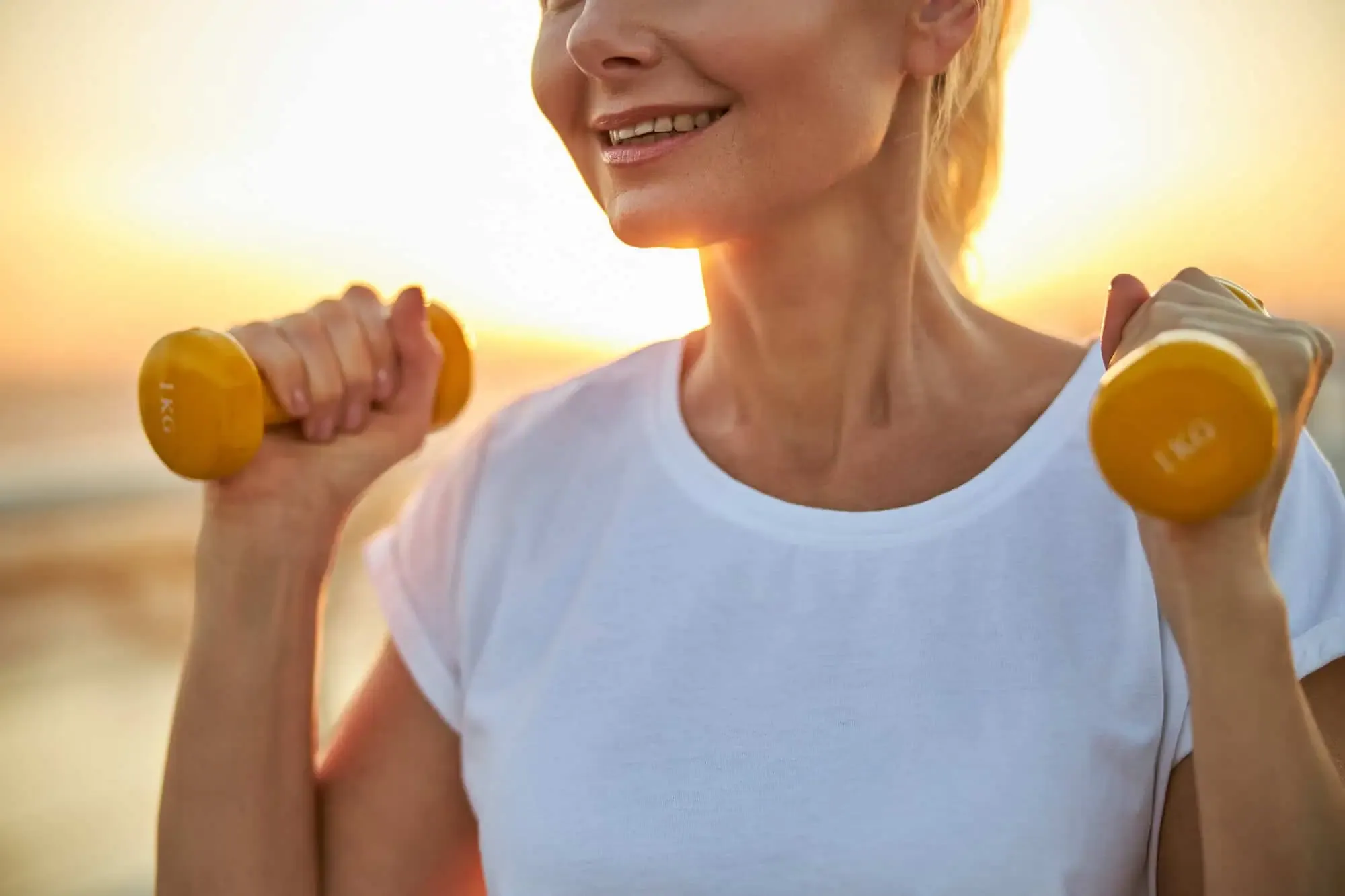 Smiling woman holding yellow 1 kg dumbbells near her shoulders at sunset.