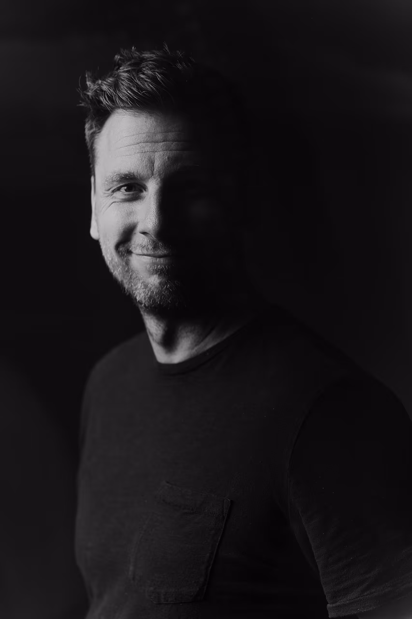 Black and white portrait of a smiling man with short hair and stubble wearing a dark shirt with a chest pocket.
