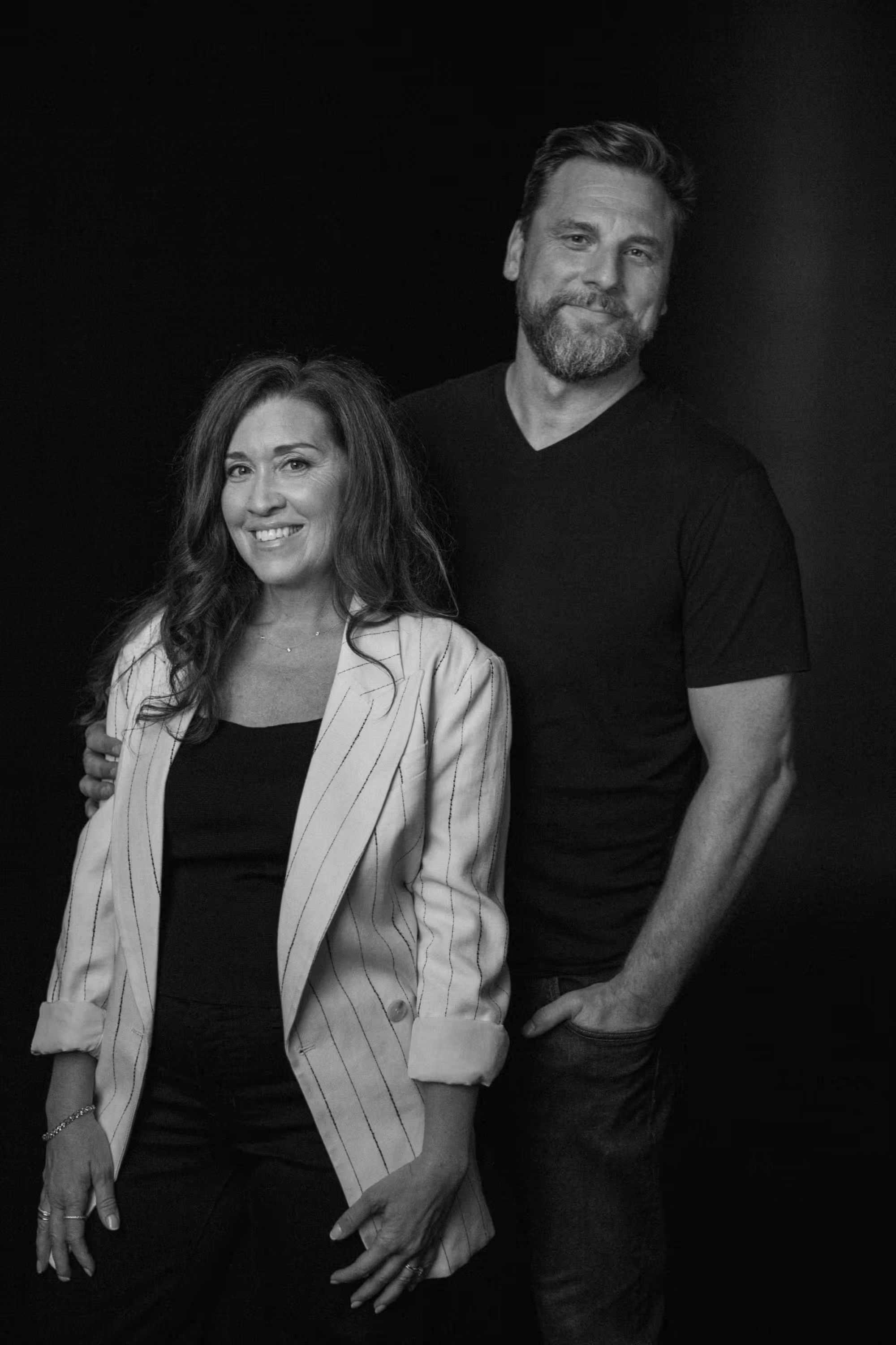 Smiling woman in a striped blazer and man in a black t-shirt posing together against a black background.