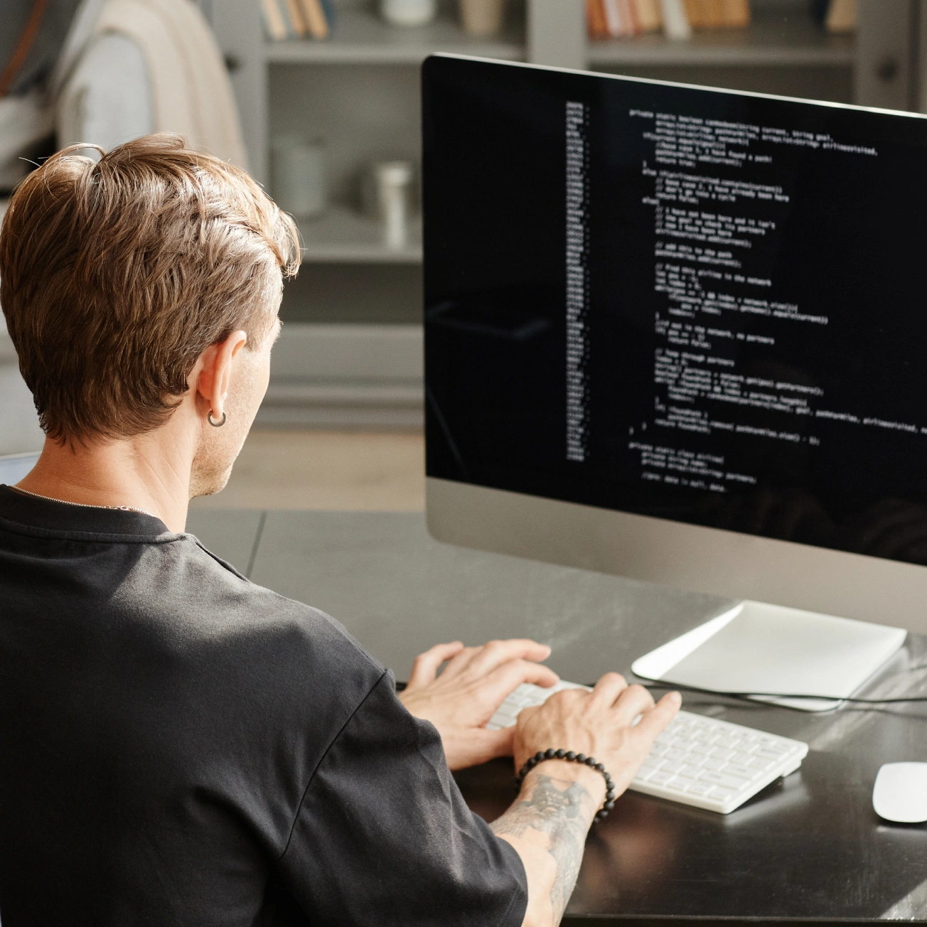 Person with tattooed arm typing on a keyboard in front of a computer screen displaying lines of code.