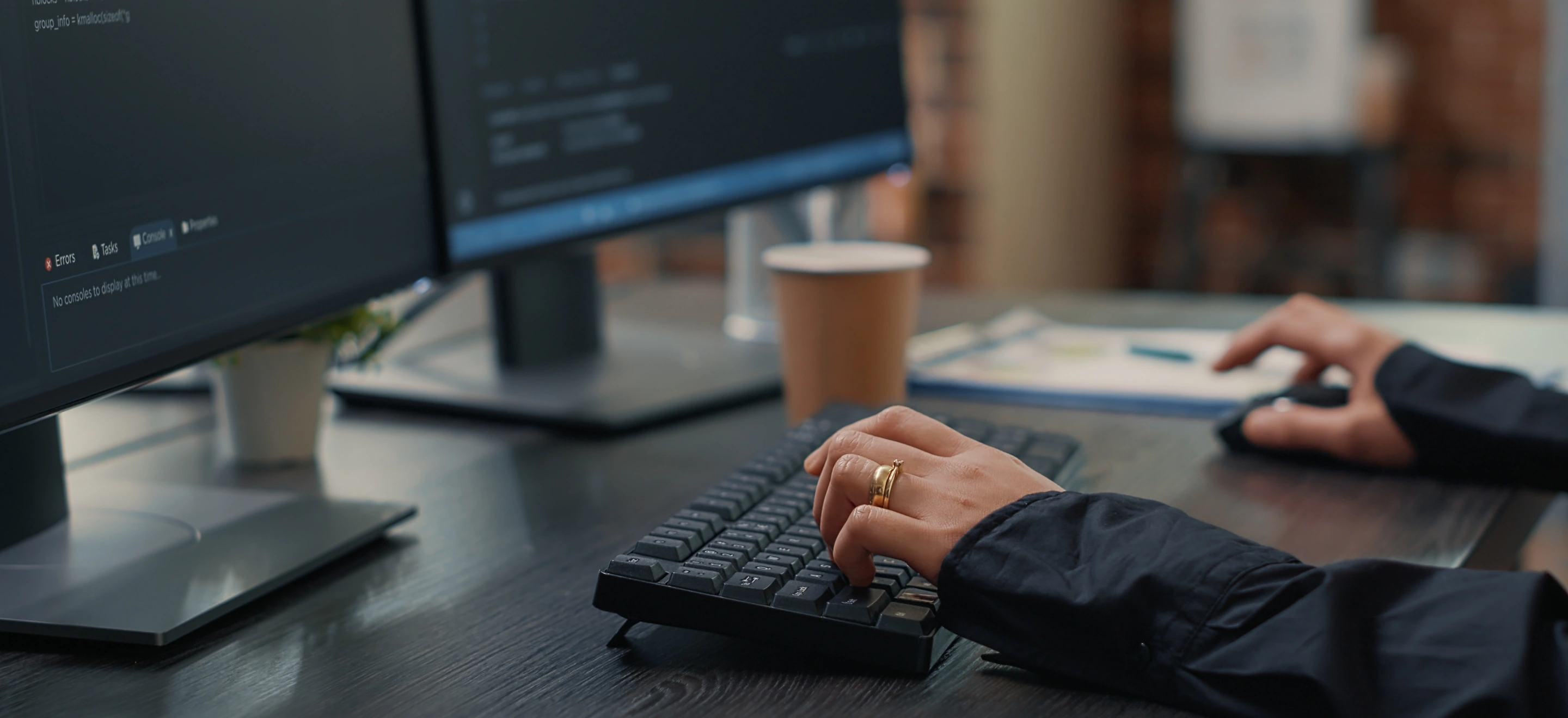 Person typing on a black keyboard with two monitors displaying code in the background and a paper cup on the desk.