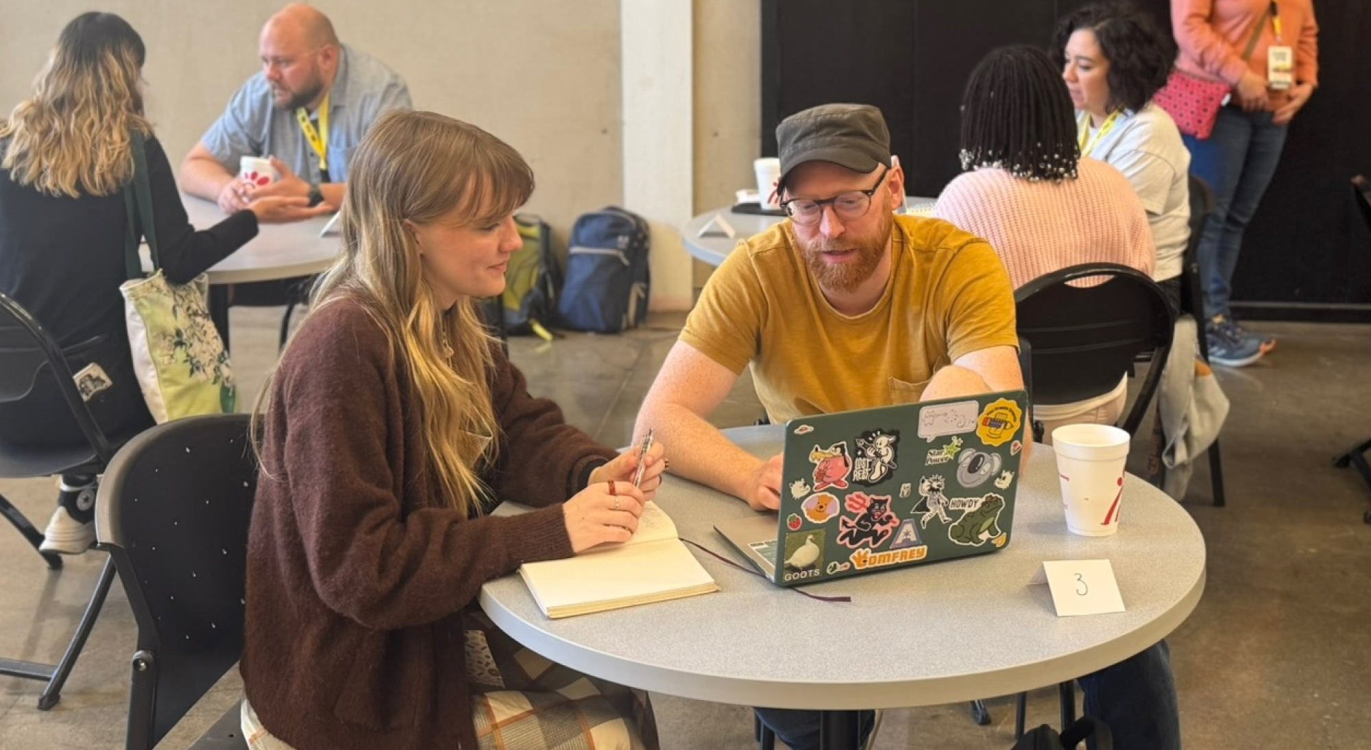 Two people sit at a round table with a laptop, talking