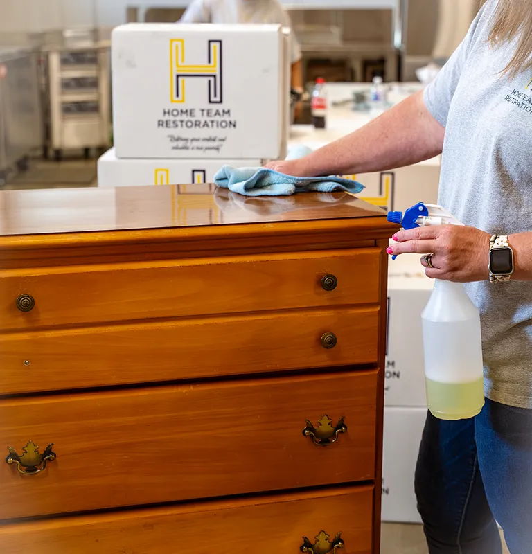 Person cleaning wooden dresser with spray bottle and cloth, Home Team Restoration boxes in background.
