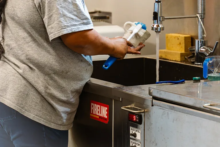 Person scrubbing a white mug with a blue cleaning brush over a kitchen sink with running water.