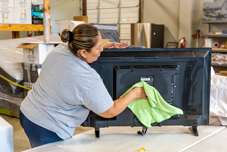 Woman cleaning the back of a flat-screen TV with a green cloth in a warehouse setting.