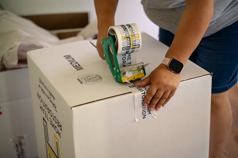 Person sealing a white medium moving box labeled Home Restoration with tape and a dispenser.