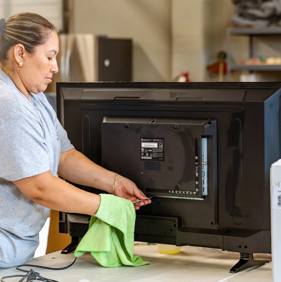 Woman in gray shirt fixing or cleaning the back of a TV with a green cloth on a table.