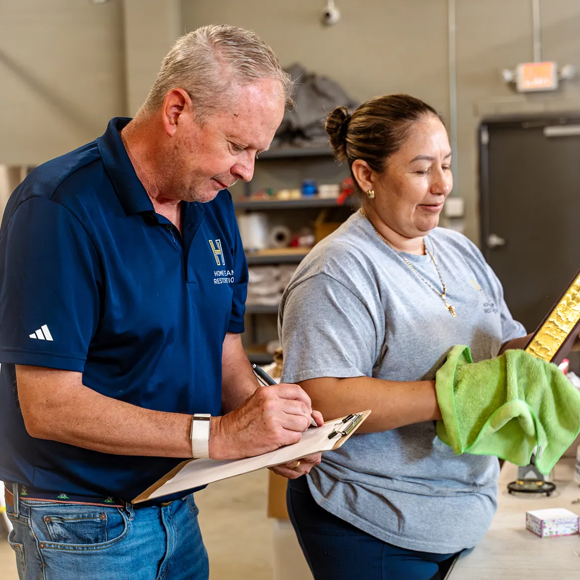 Man in blue polo writing on clipboard next to woman holding a golden framed object with a green cloth.