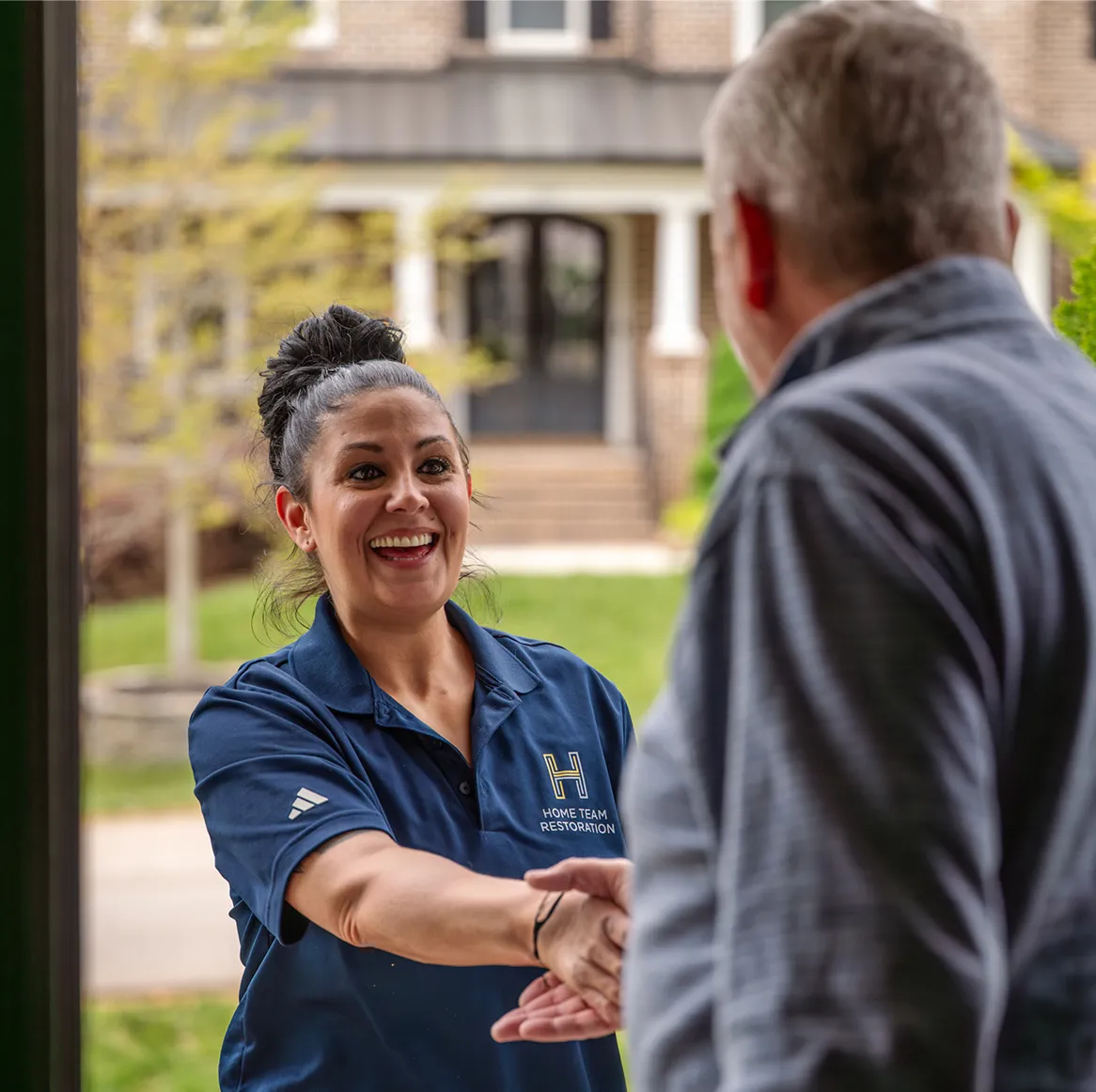 Smiling woman in a blue Home Team Restoration shirt shaking hands with a man in front of a house.