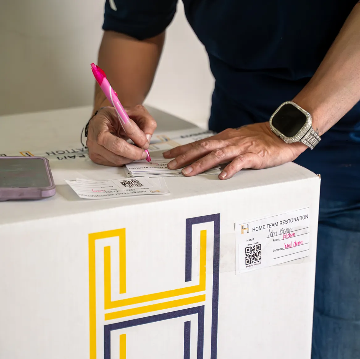 Person with tattooed arms writing on label attached to a box labeled Home Team Restoration.