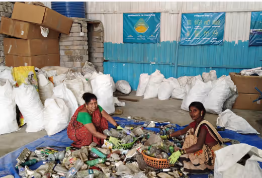 Two women sorting recyclable plastic bottles on a tarp inside a recycling facility with sacks and boxes in the background.