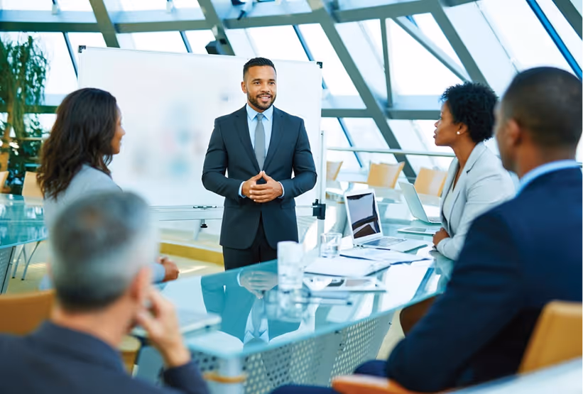 Businessman in a suit giving a presentation to colleagues seated around a glass conference table in a modern office.