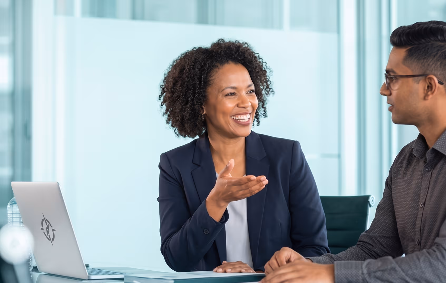 Two professionals, a woman and a man, engaged in a friendly conversation at a modern office desk with a laptop.