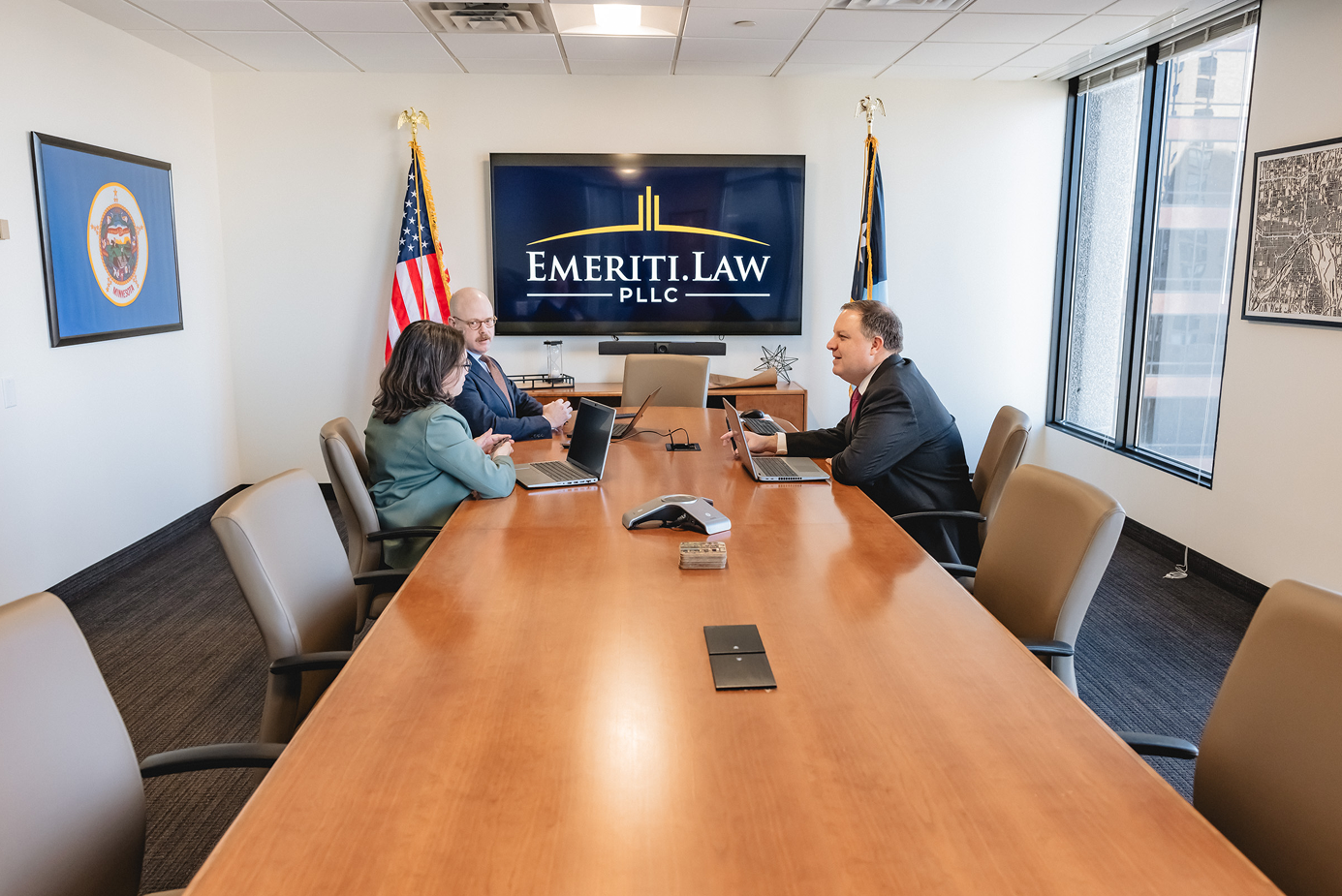 Ryan R. Wood, Wynne P. Kelly and Elizabeth seated around a conference table with laptops in a law office meeting room with Emeriti Law PLLC logo on a screen.