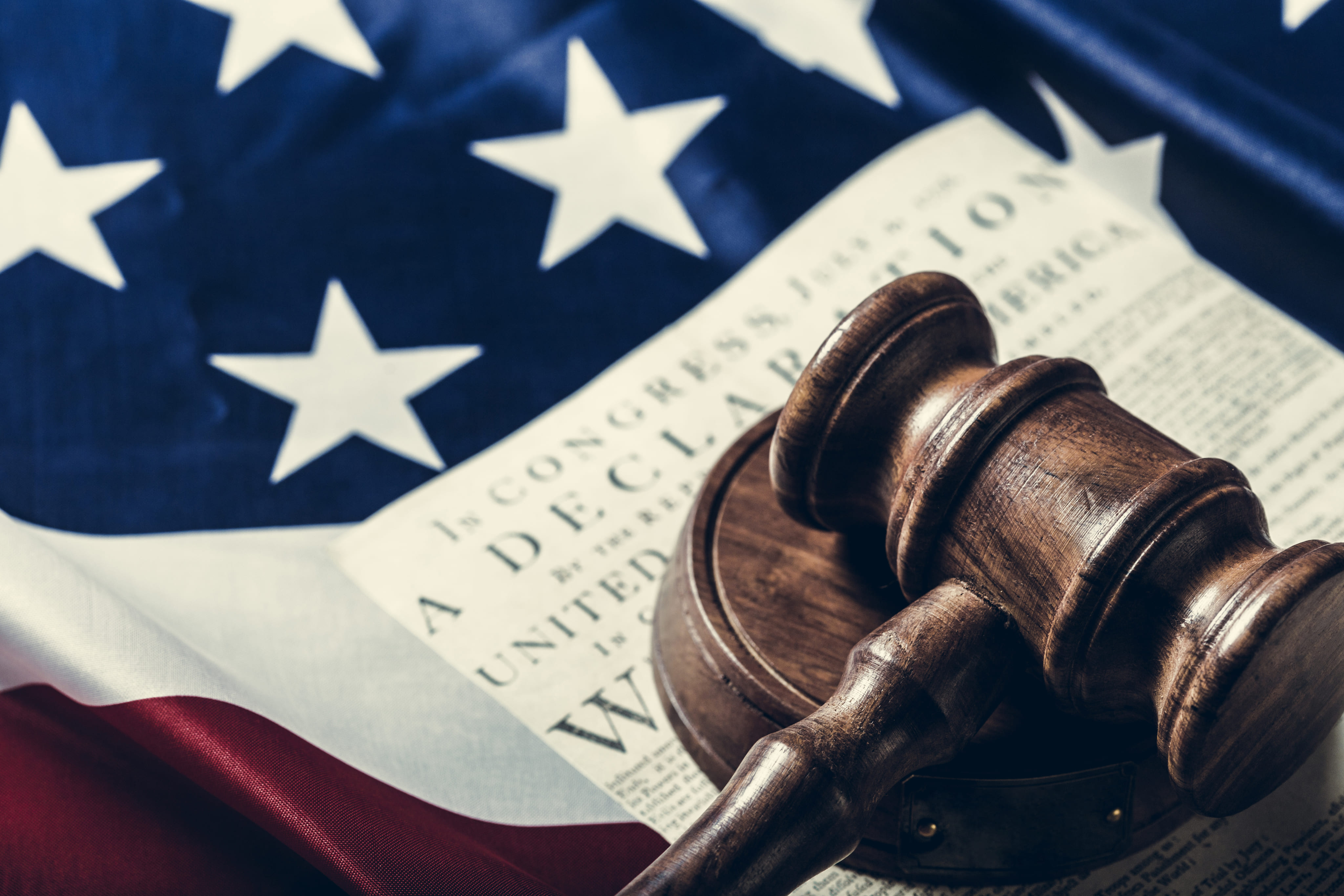 Wooden judge's gavel resting on a copy of the Declaration of Independence over a folded U.S. flag.
