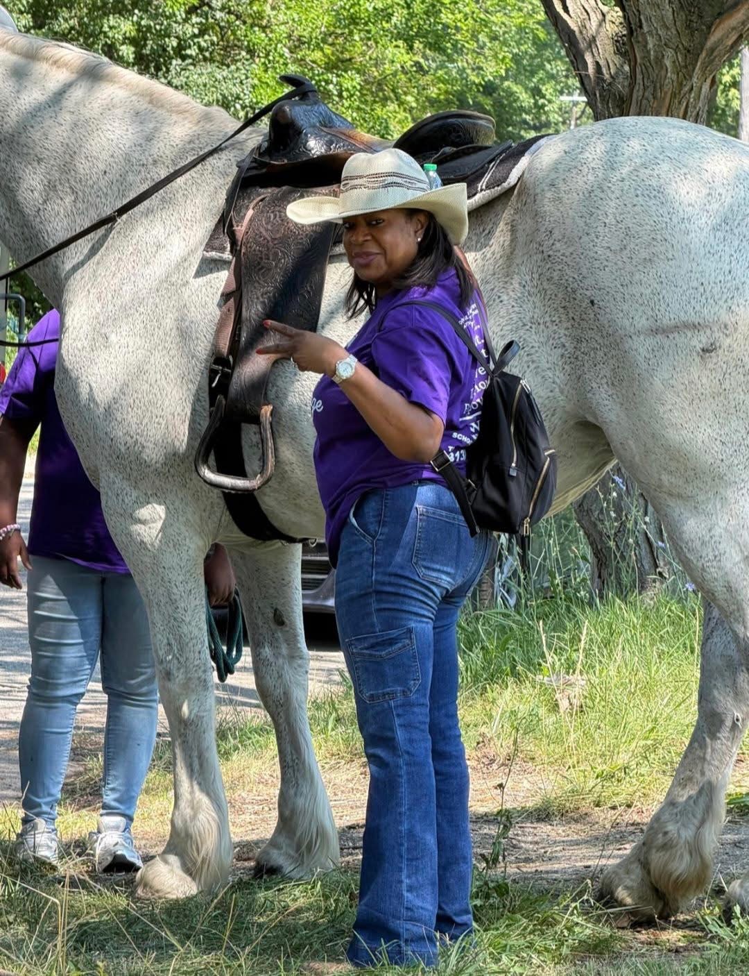 The side profile of Dr. Martine Francois is showcased as she stands on the side of Bell, a white female Percheron. Dr. Francois has her cowgirl attire on with her purple Queen Carriage Horse Rides shirt.
