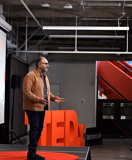 Man in a brown jacket speaking on stage at a TEDx event with industrial background.