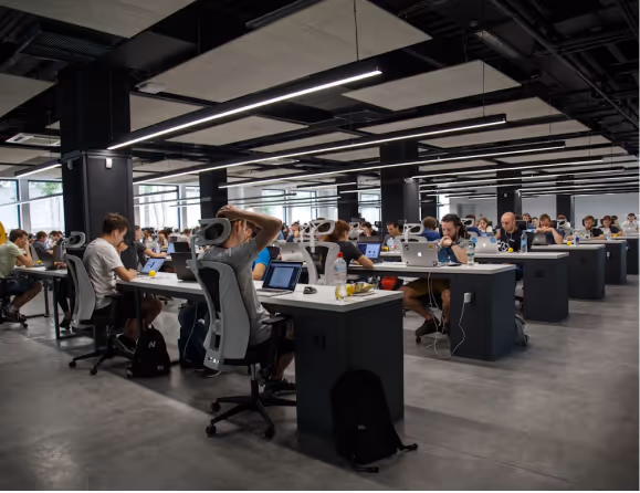 Large modern workspace with rows of people working on laptops at desks in an open office environment.