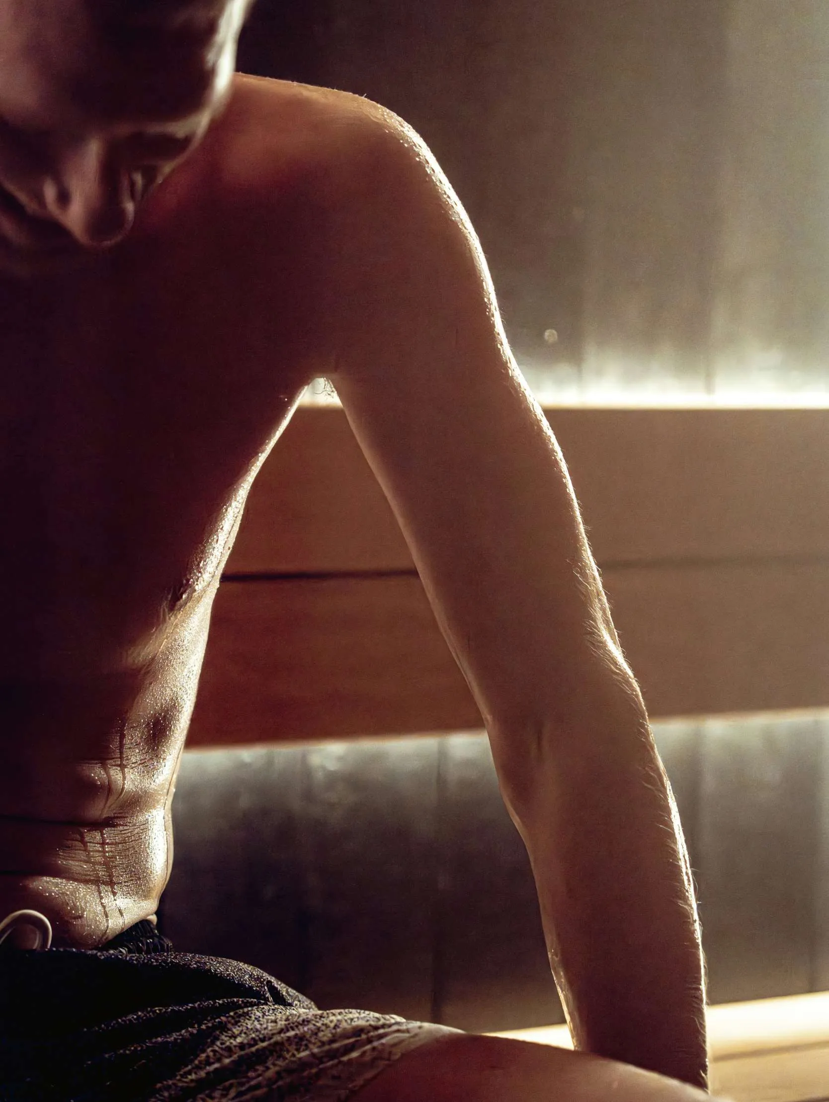 Close-up of a shirtless man sitting with visible sweat on his arm and torso in warm light.