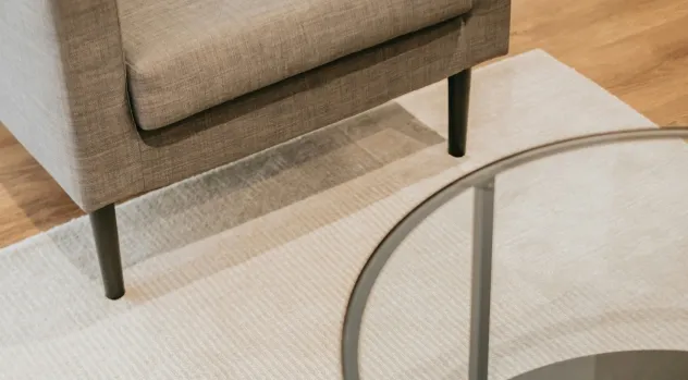 Close-up of a beige fabric sofa leg on a textured light rug beside a glass and metal round coffee table.