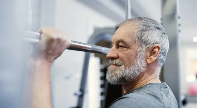 Older man with gray hair and beard lifting a barbell at a gym.