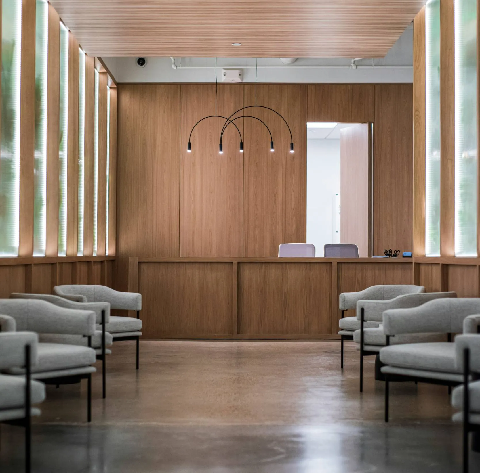 Modern waiting area with rows of gray cushioned chairs facing a wooden reception desk under minimalist black pendant lights.