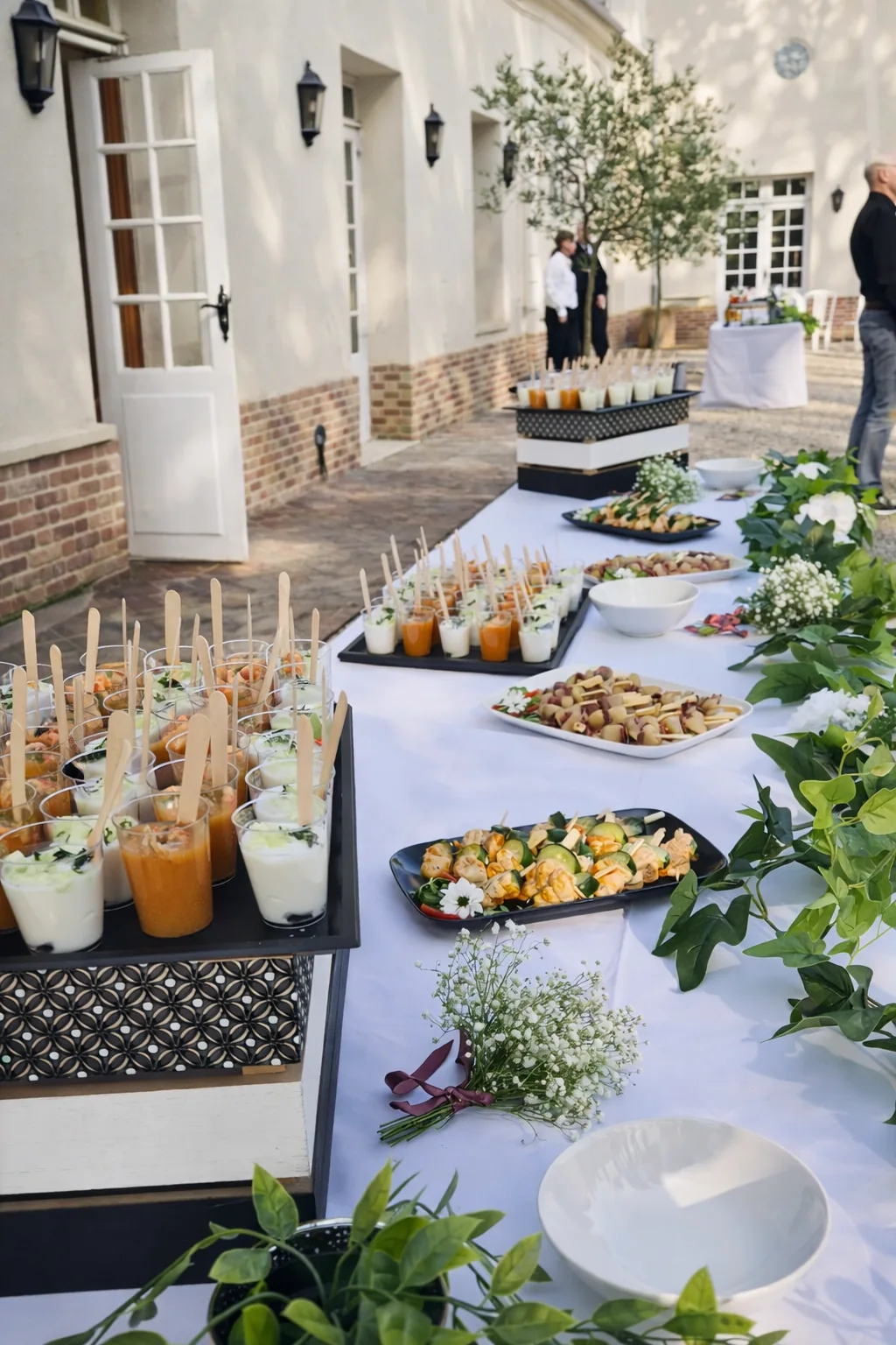 Buffet cocktail dinatoire avec verrines colorées, brochettes et mignardises présentés sur longue table en cour intérieure avec olivier en pot pour une réception élégante.