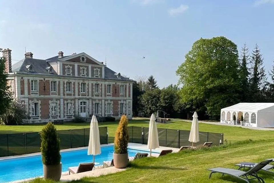 Château en briques rouges avec piscine paysagée, parasols fermés et chapiteau blanc de réception dans un parc verdoyant.