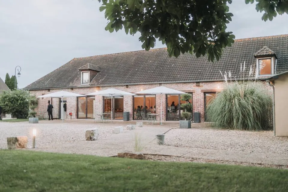 Grange réceptive en briques rouges avec parasols blancs et cour gravillonnée — lieu de réception champêtre.