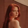 Portrait intime d'une jeune femme rousse aux longs cheveux ondules, regard fixe, lumiere chaude sur fond brun pour temoignage client.