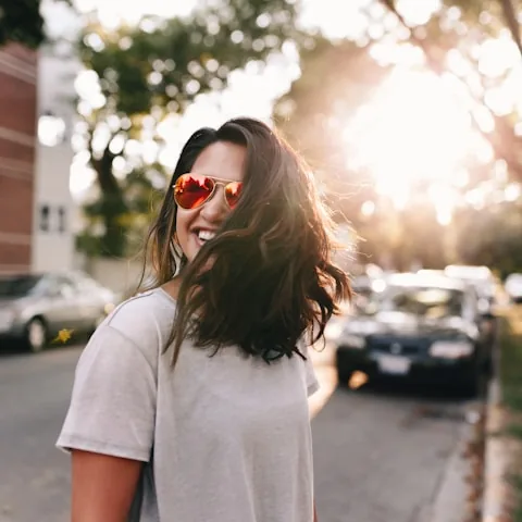 Portrait souriant d'une femme avec lunettes de soleil reflechissantes, cheveux dans le vent, dans une rue ensoleillee en contre-jour.