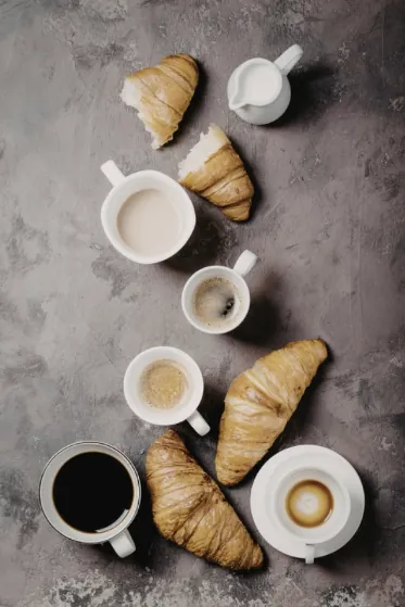 Composition de petit-déjeuner vue de dessus avec croissants dorés, tasses de café noir et cappuccino disposés sur fond gris ardoise.