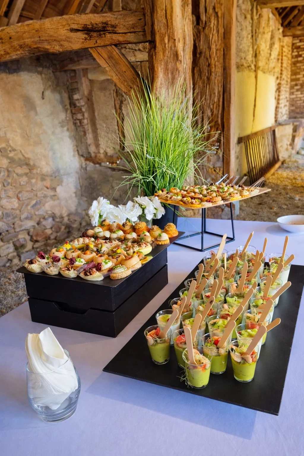 Buffet cocktail dinatoire dans une grange avec verrines de guacamole, brochettes et mignardises sucrées présentés sur plateaux ardoise et bois.