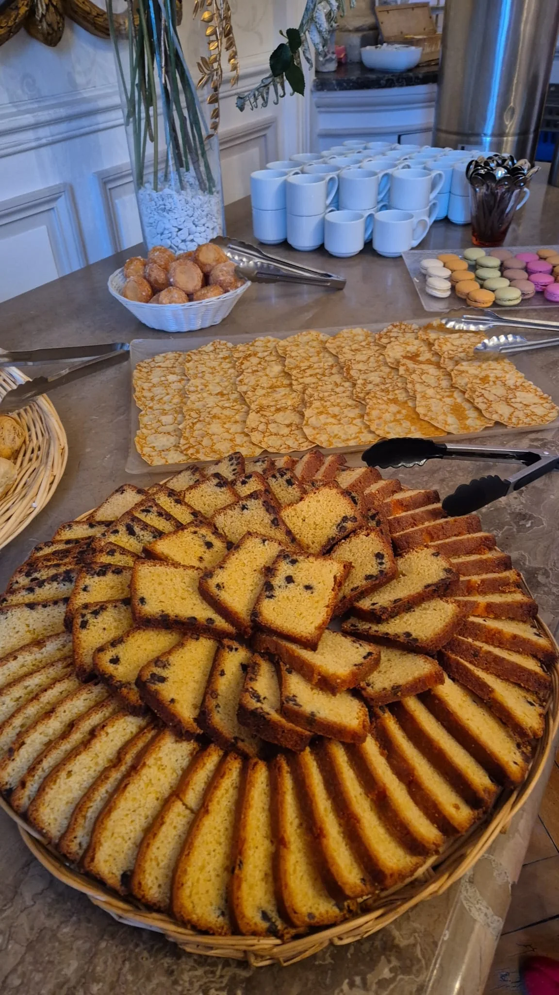 Buffet de brunch avec un grand cake marbre tranche en eventail dans un panier osier, tuiles aux amandes, macarons et cafes en arriere-plan.