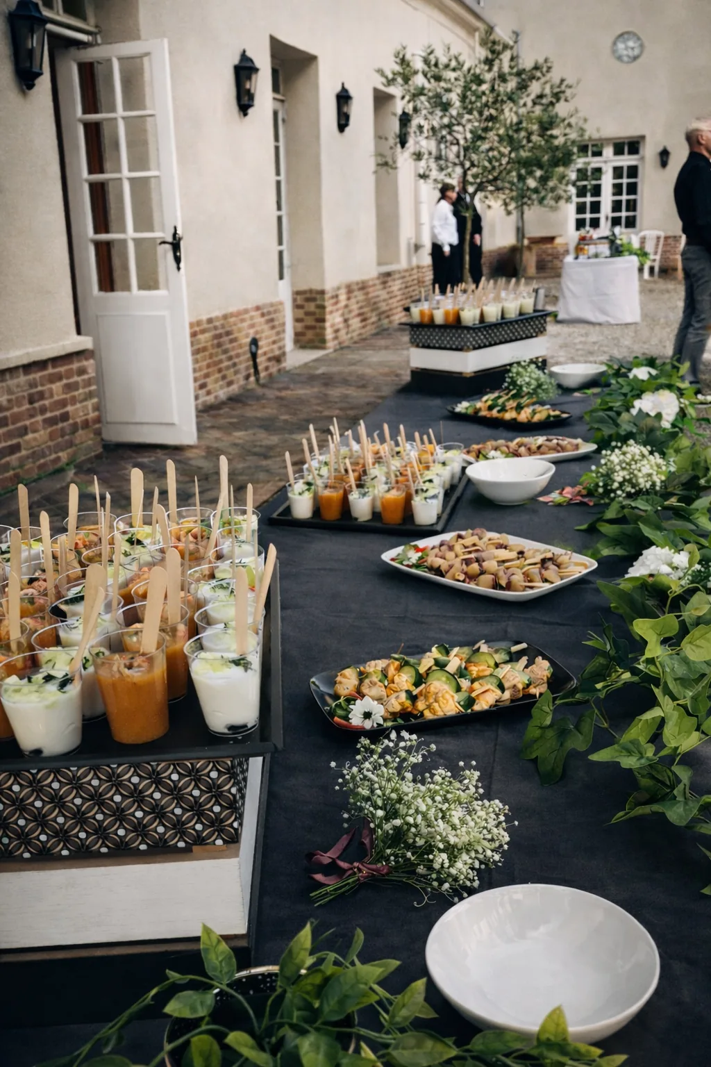 Buffet cocktail dinatoire avec nappe noire présentant verrines, brochettes et mignardises décorées de gypsophiles en cour pavée pour une réception professionnelle.