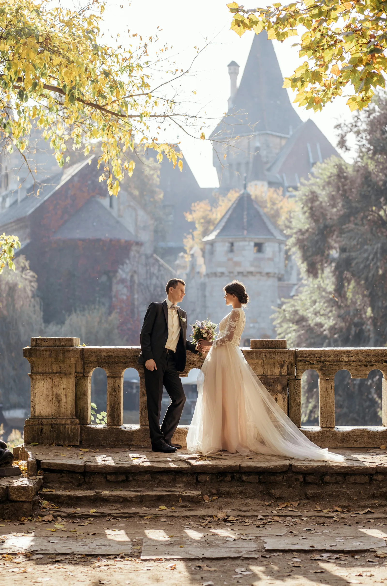 Couple de mariés sur un pont en pierre devant un château automnal, mariée en robe princesse et marié en costume sombre.