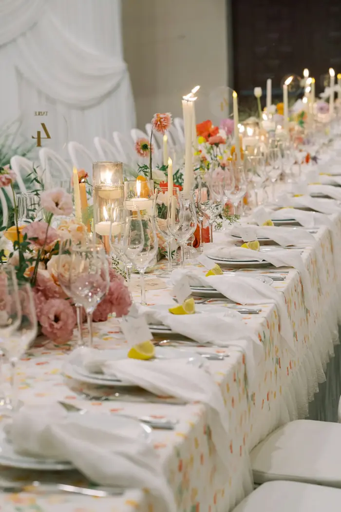 Longue table de mariage dressée avec nappe fleurie pastel, hautes bougies, citrons coupés et compositions florales abondantes.