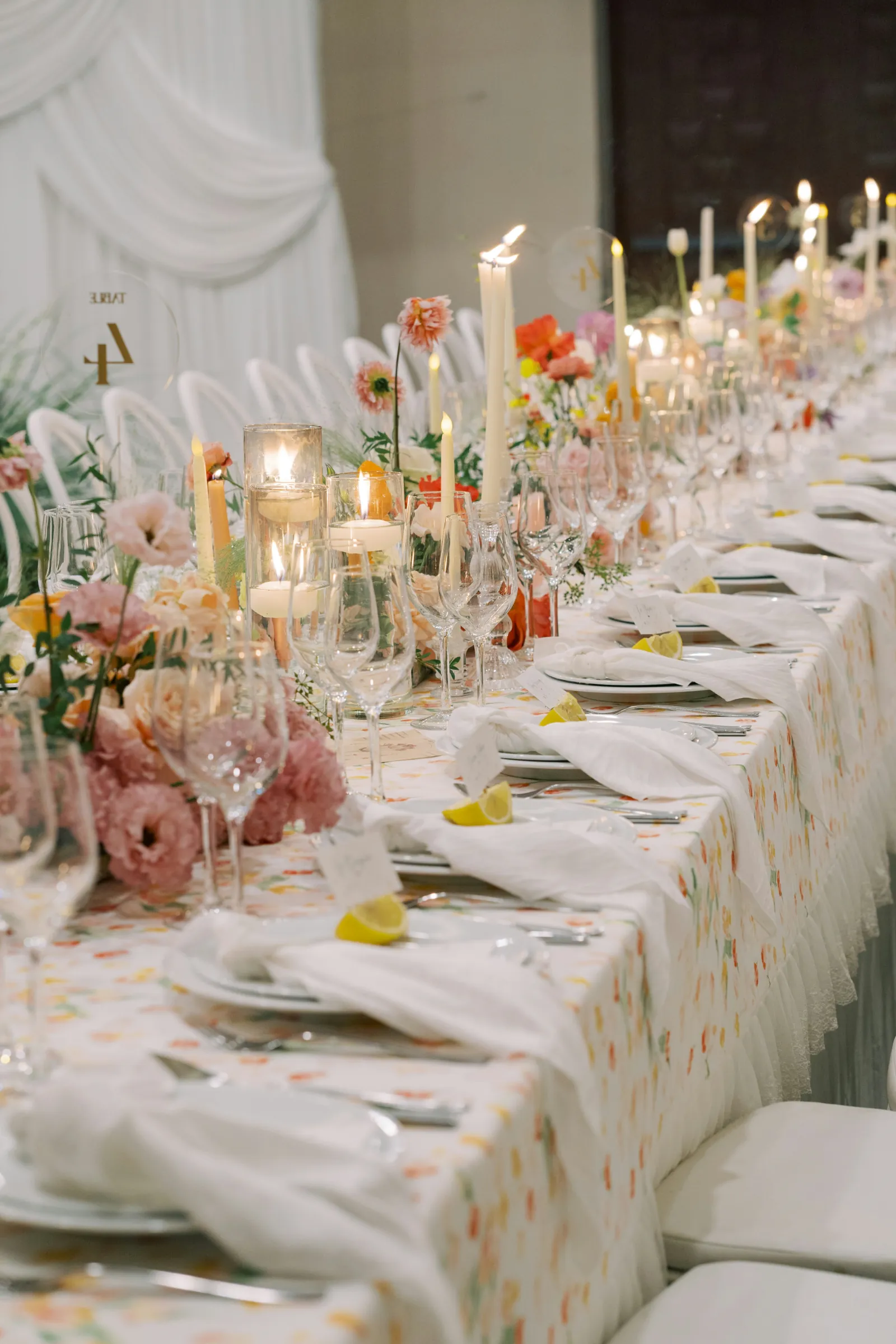 Longue table de mariage dressée avec nappe fleurie pastel, hautes bougies, citrons coupés et compositions florales abondantes.