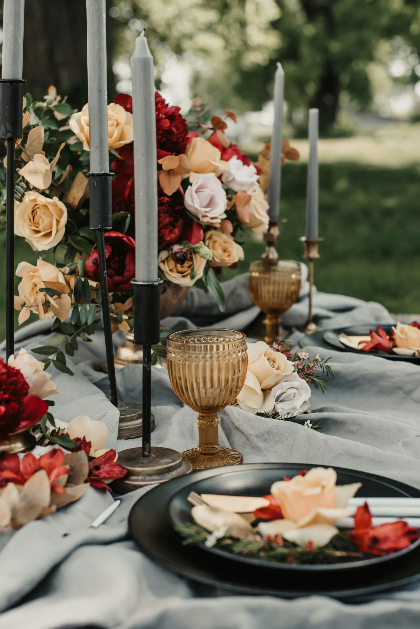 Détail décoration de table de mariage en jardin : bougies grises hautes, verre vintage ambré, composition florale rouge et crème.