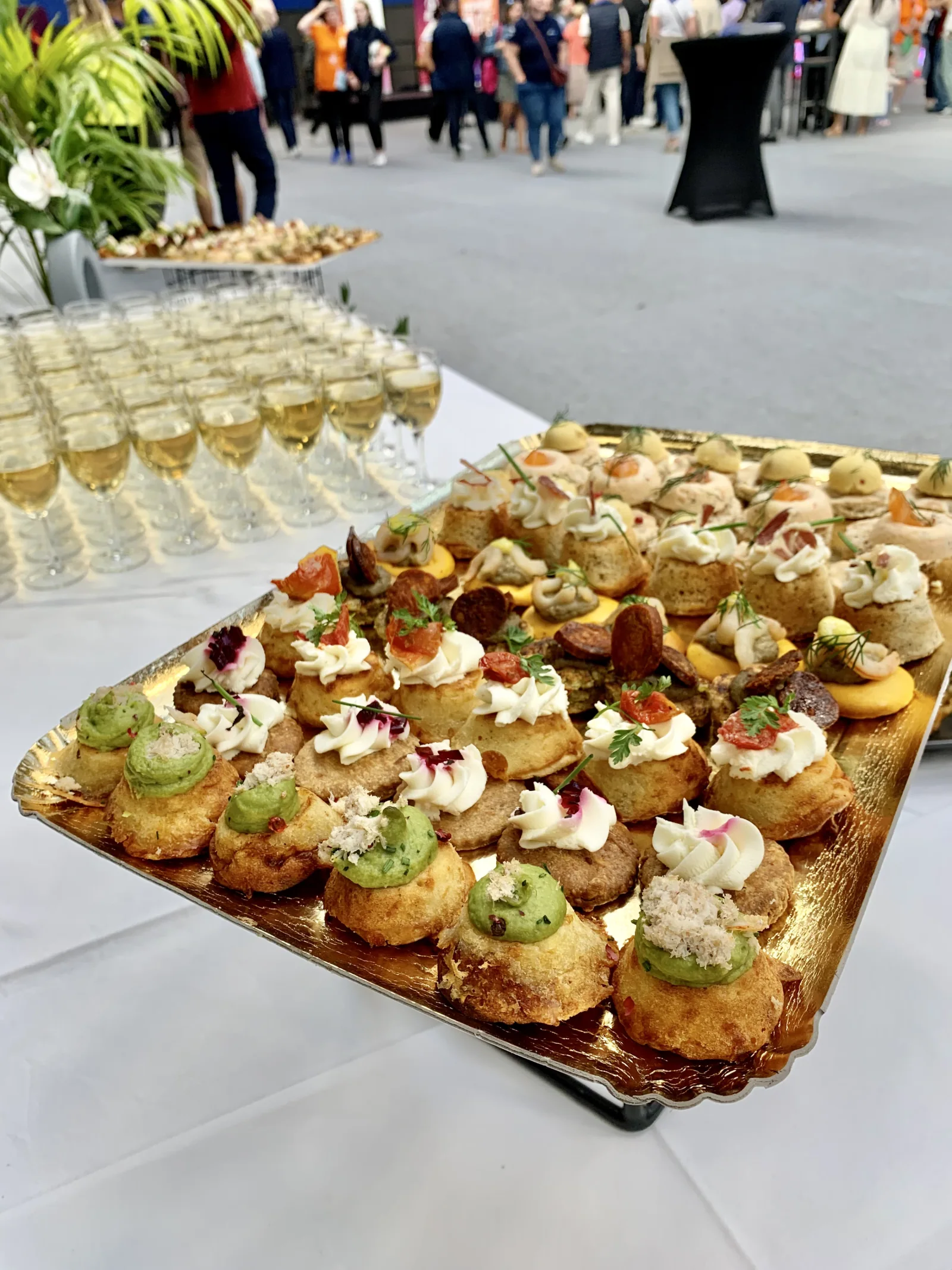 Plateau doré de mini-choux salés garnis de mousses colorées en arrière-plan d'un cocktail événementiel avec coupes de champagne alignées.